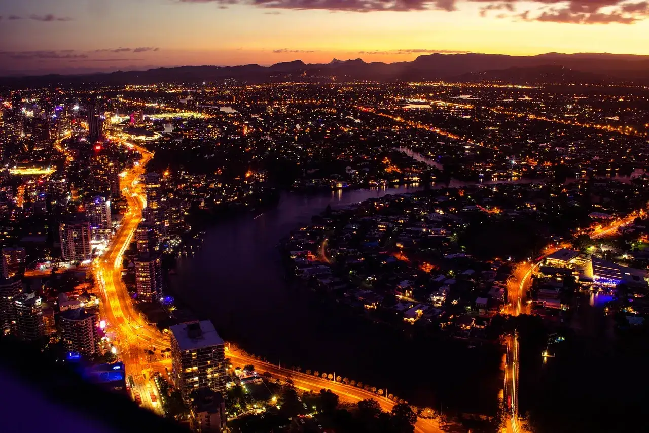Vue a&eacute;rienne d'une ville illumin&eacute;e au cr&eacute;puscule, avec une rivi&egrave;re sinueuse traversant le paysage urbain. Les lumi&egrave;res des b&acirc;timents et des rues cr&eacute;ent un spectacle scintillant.