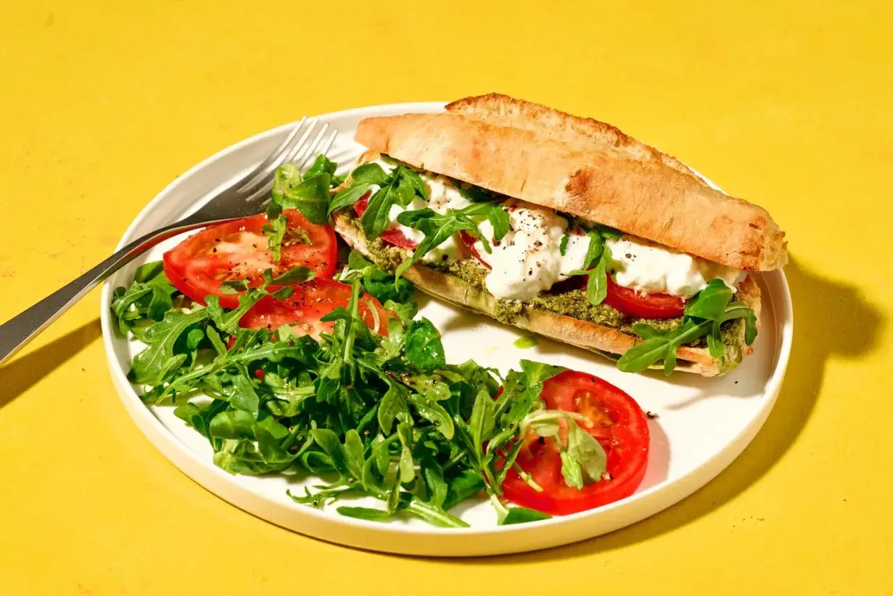 Un d&eacute;licieux sandwich burrata garni de pesto, tomates et roquette, servi avec une salade fra&icirc;che.