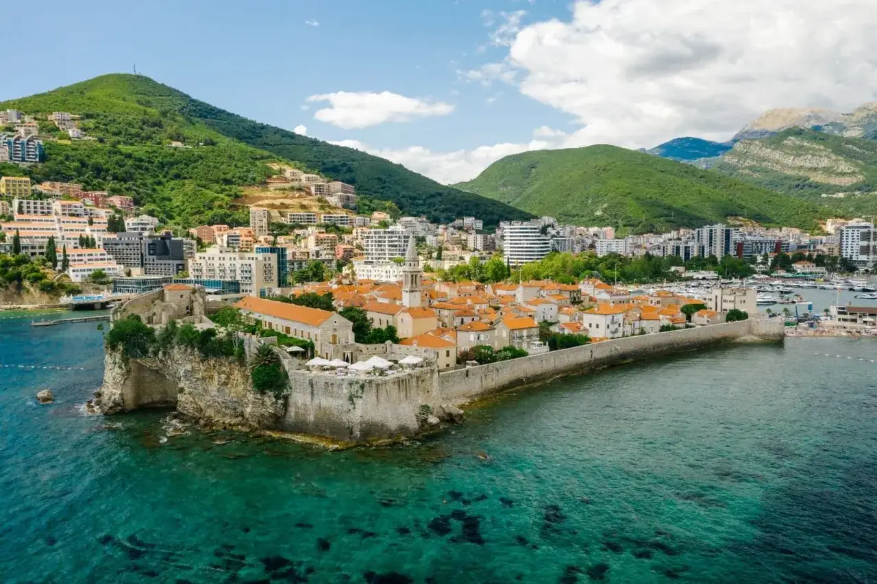Widok na starówkę Budvy z murami obronnymi i turkusową wodą. W tle zielone wzgórza i nowoczesne budynki, idealne miejsce na budva plaże.