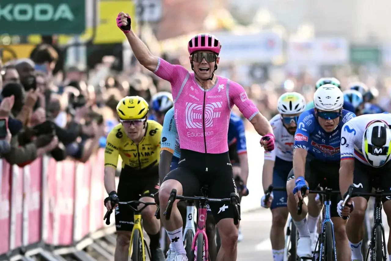 Luke Lamperti celebrates a victory, raising his fist in triumph as he crosses the finish line in a pink cycling jersey, surrounded by other racers and a cheering crowd.