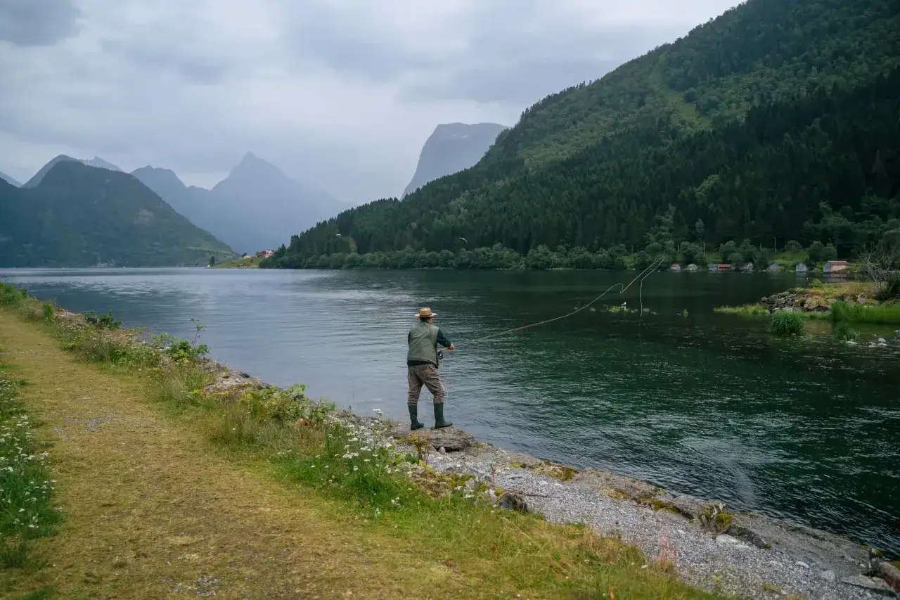 Ein Mann angelt in Norwegen an einem ruhigen Fjord. Gr&uuml;ne Berge und kleine H&auml;user s&auml;umen das Ufer.