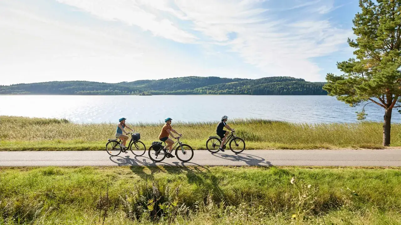 Tre personer cyklar l&auml;ngs en sj&ouml; p&aring; en asfalterad v&auml;g, en idyllisk scen som visar upp Sveriges vackra cykelleder.