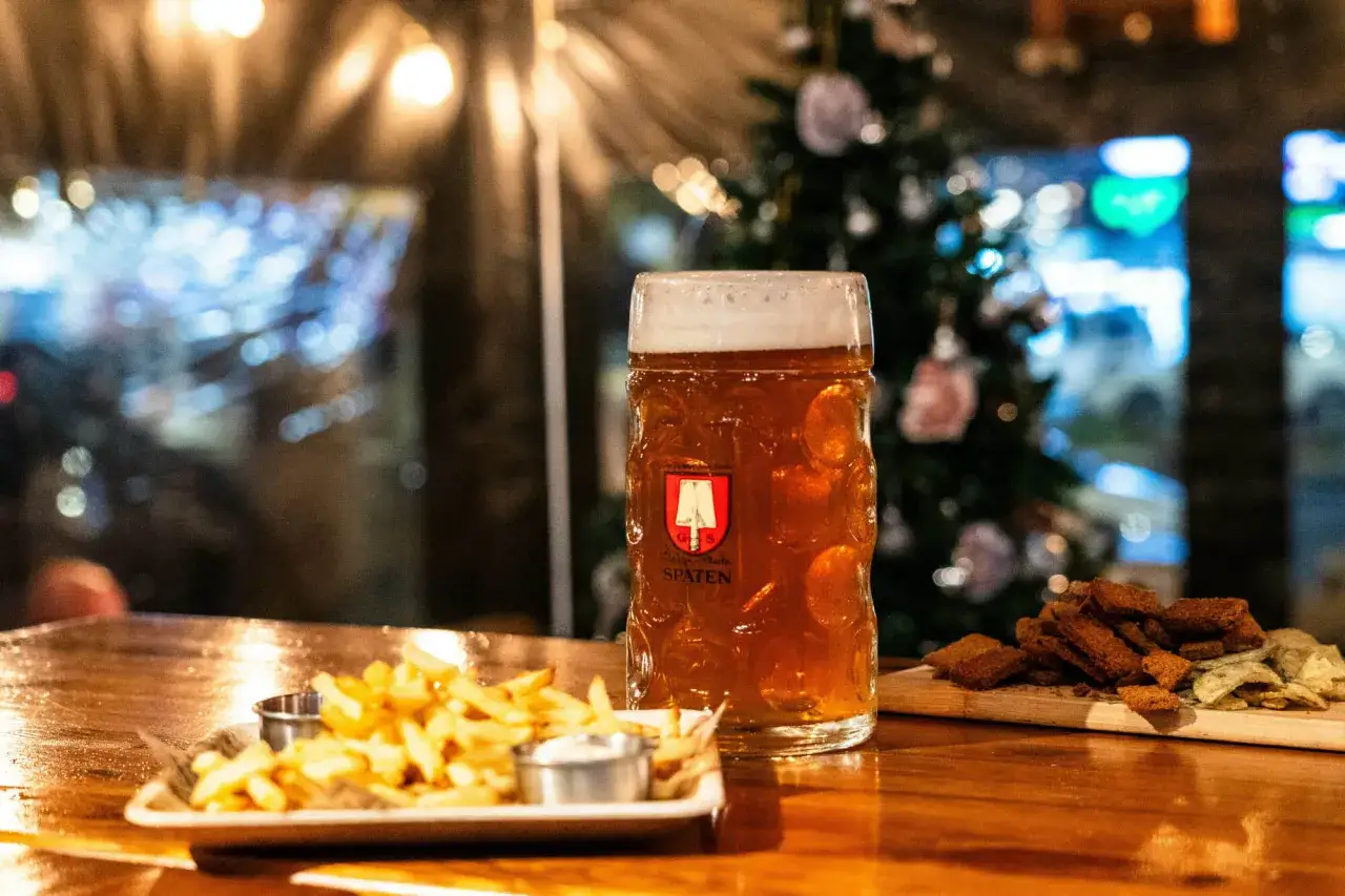 Une chope de bi&egrave;re ambr&eacute;e, des frites et des croutons sur une table en bois, avec un sapin de No&euml;l d&eacute;cor&eacute; en arri&egrave;re-plan. Parfait pour une soir&eacute;e bi&egrave;re chaude No&euml;l.