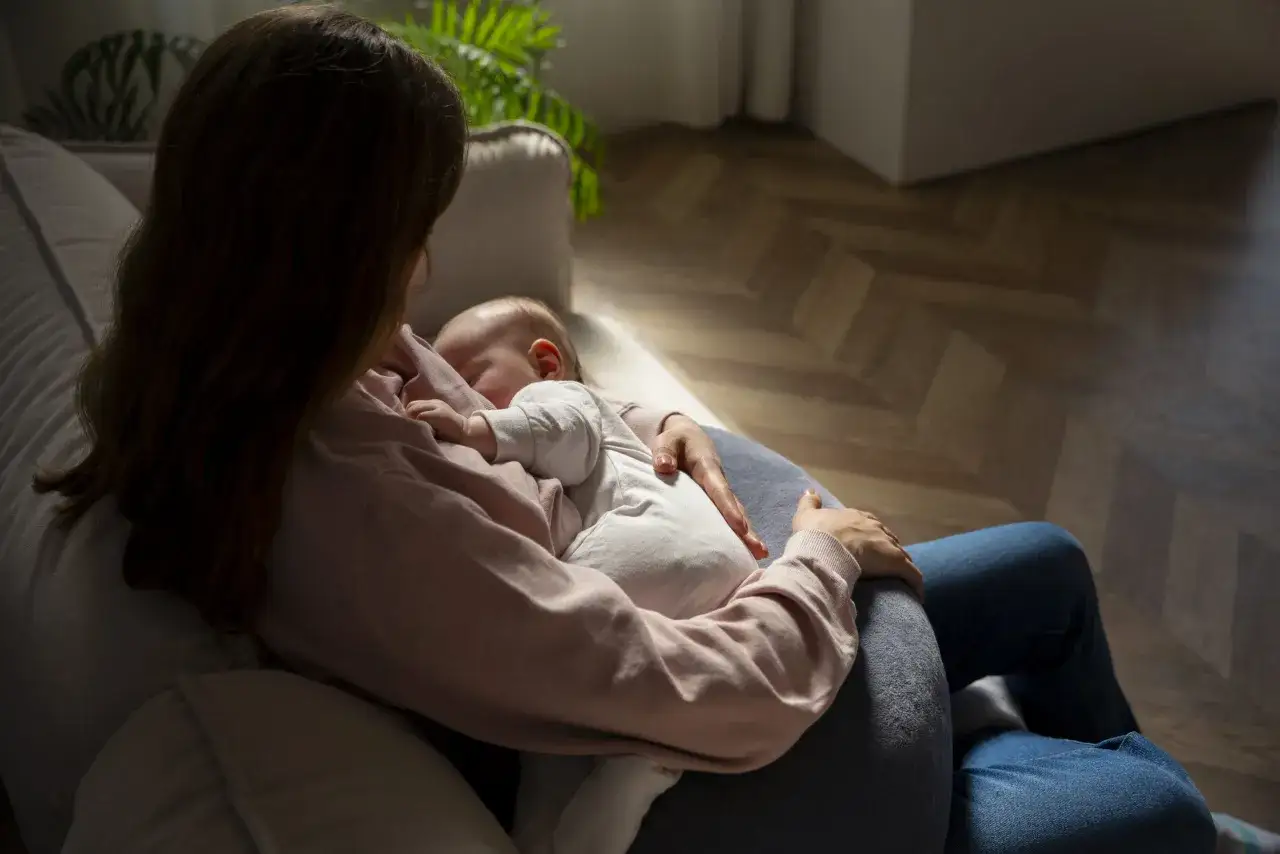 Kobieta karmi piersią dziecko, czując się przytłoczona. Ciemne światło podkreśla jej smutek, być może objaw depresji poporodowej.