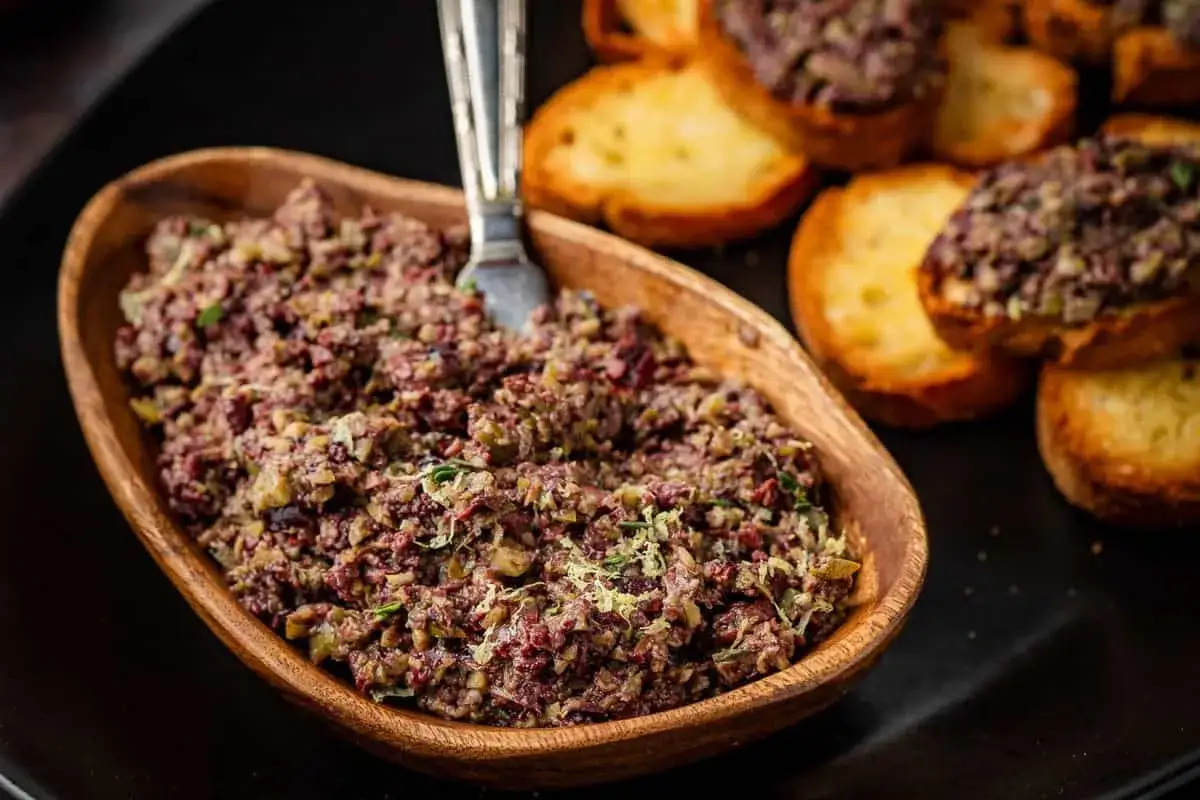 A rustic wooden bowl overflows with a chunky tapenade, a delicious spread made from olives, capers, and herbs, served with toasted baguette slices.
