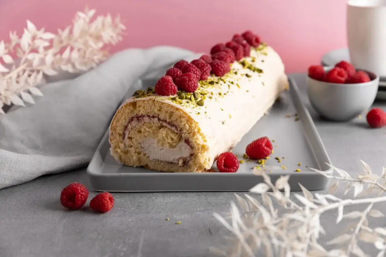 Eine k&ouml;stliche Biskuitrolle mit Himbeeren, verziert mit Pistazien und frischen Beeren, wartet darauf, genossen zu werden.