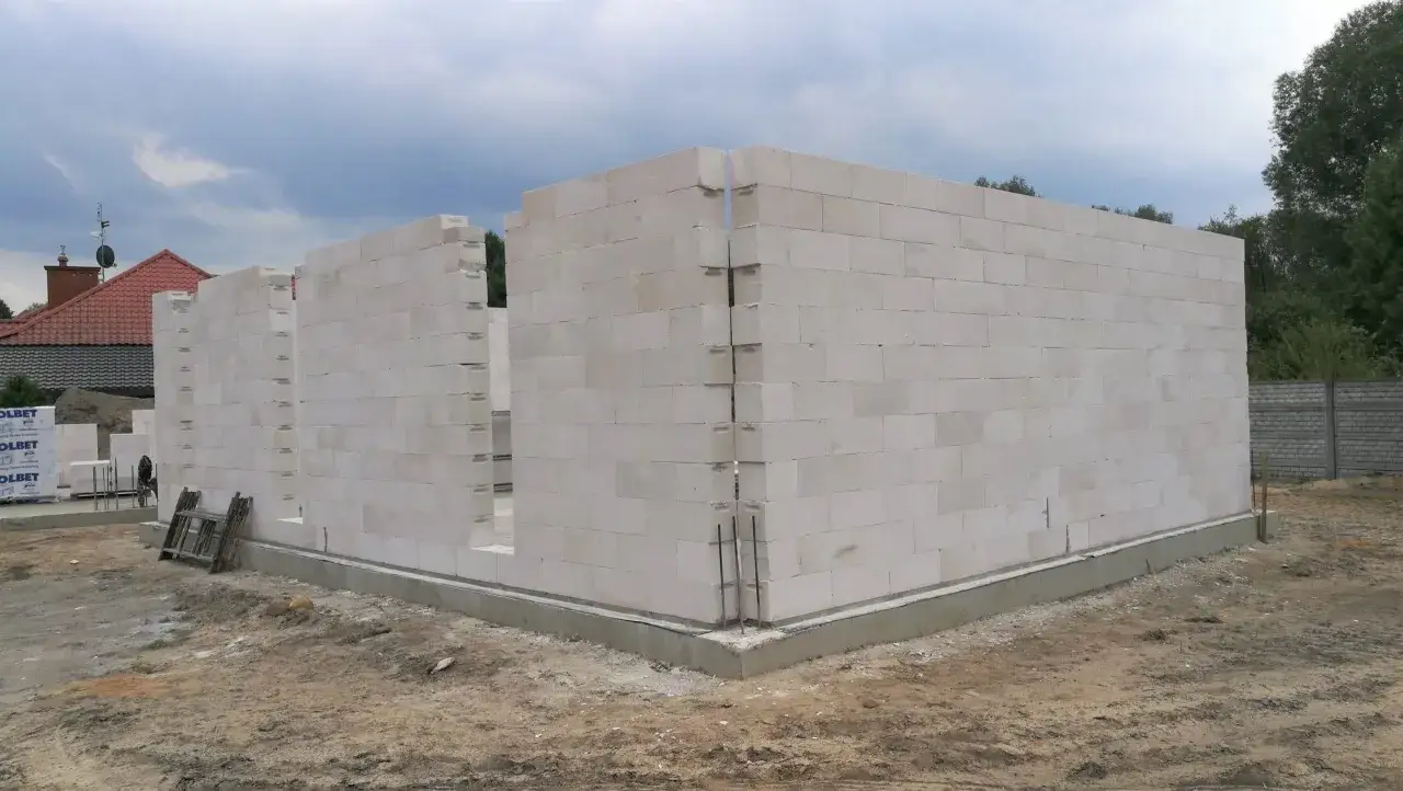 Budowa domu z pustaków z betonu komórkowego. Ściany wznoszą się na fundamencie, w tle widać dach sąsiedniego budynku.