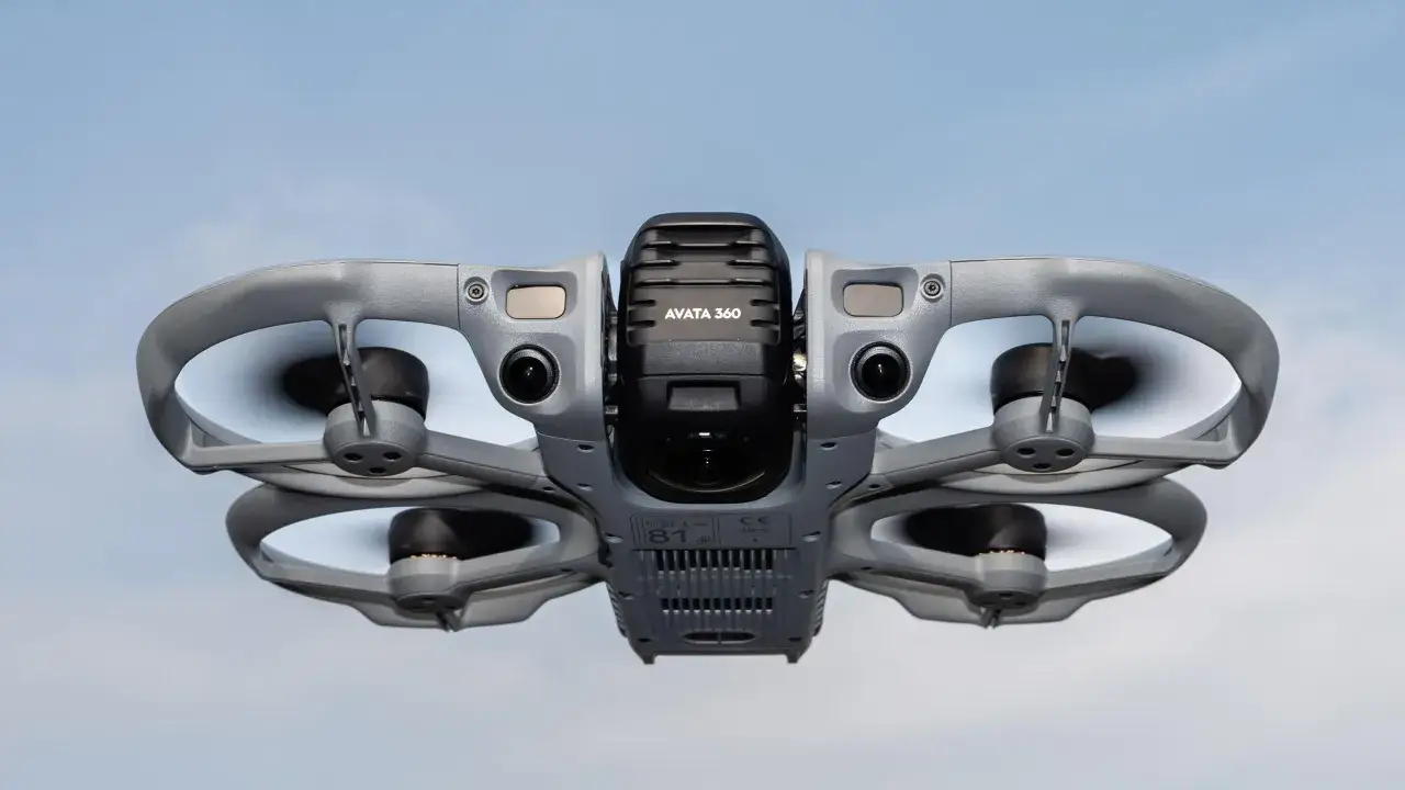 Szary dron DJI Avata 3 z kamerą i śmigłami unosi się na tle błękitnego nieba.