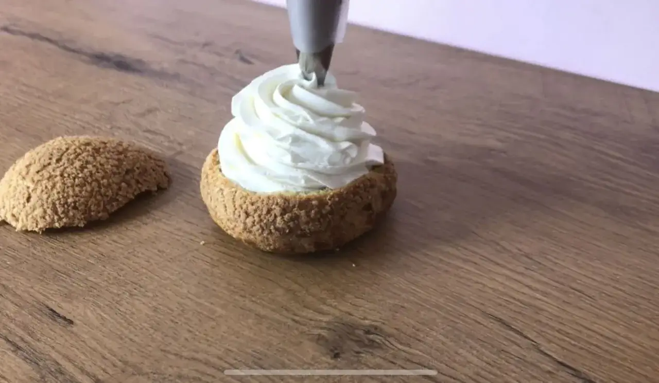 Une poche à douille dépose une généreuse crème chantilly sur un chou craquelin. Un autre chou est posé à côté.