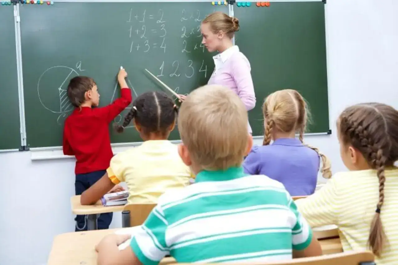Nauczycielka tłumaczy matematykę, a chłopiec pisze na tablicy. Dzieci siedzą w ławkach, słuchając lekcji. To piękny wiersz o szkole.