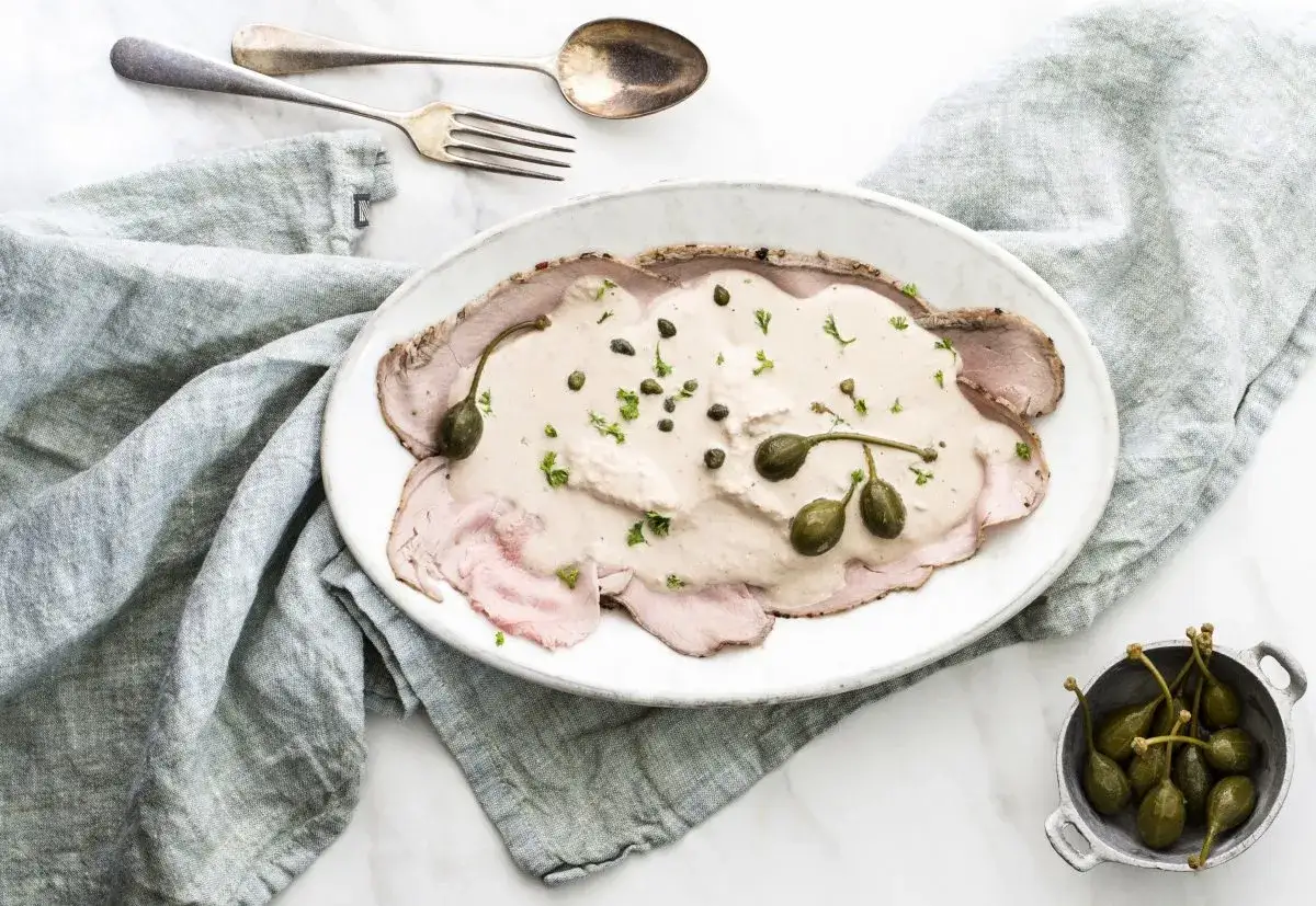 Eleganckie vitello tonnato z kremowym sosem, kaparami i natką pietruszki na białym talerzu. Obok miseczka z kaparami.
