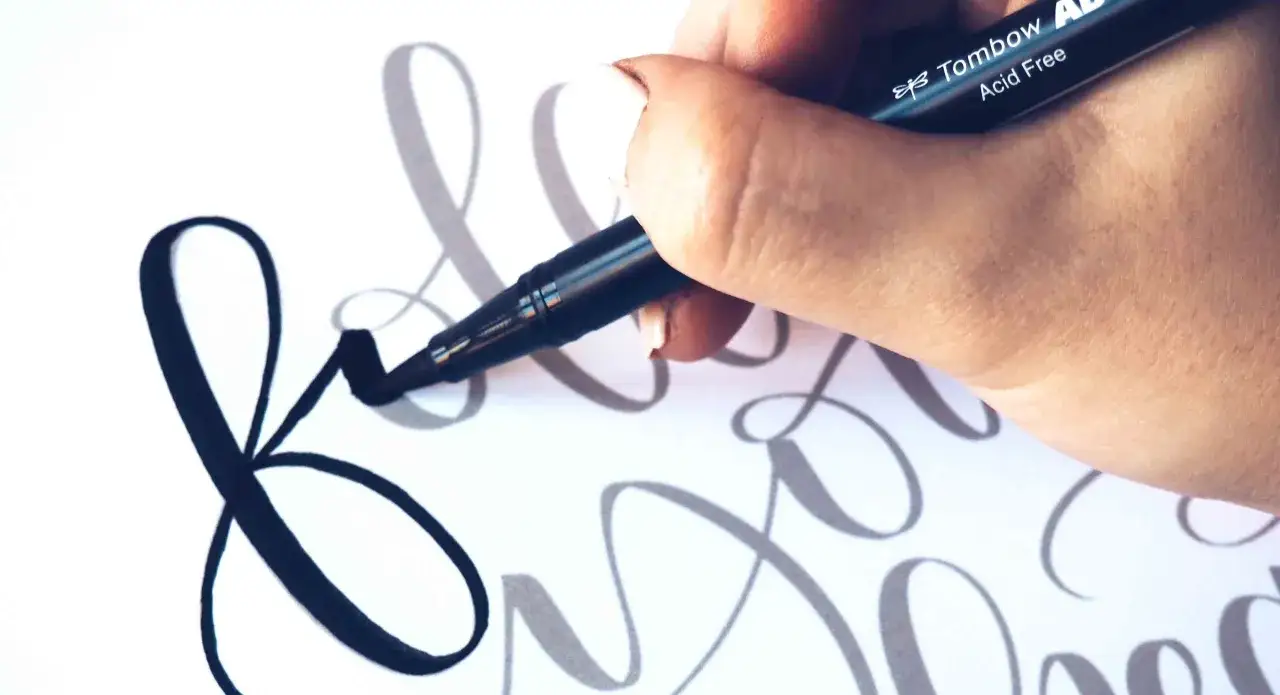 Mano practicando cómo hacer lettering con un rotulador Tombow sobre papel blanco, creando letras cursivas elegantes.