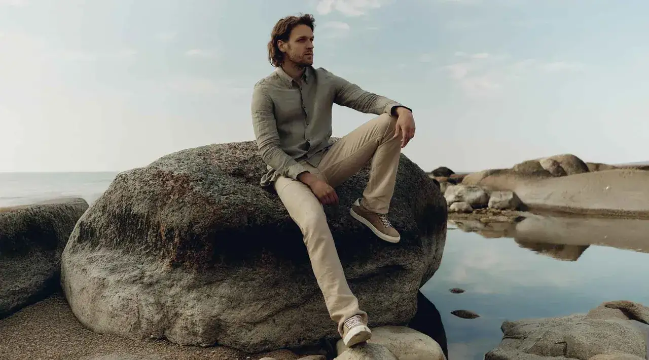 Man i linnebyxor outfit sitter på en sten vid havet.