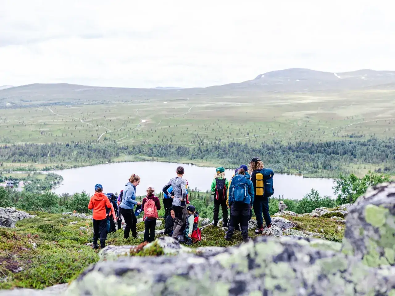 En grupp m&auml;nniskor, inklusive barn, deltar i guidade vandringar i ett vackert fj&auml;llandskap med en sj&ouml; i bakgrunden.