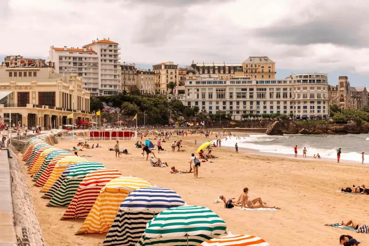 Playa con sombrillas a rayas y gente disfrutando del sol. Perfecto para aprender c&oacute;mo se dice verano en franc&eacute;s.
