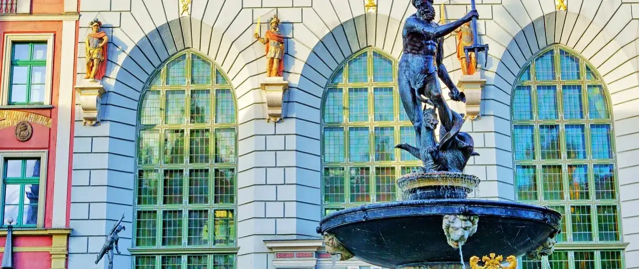 Fontanna Neptuna na tle zabytkowej gdanskiej architektury z rzeźbami i wielkimi oknami.