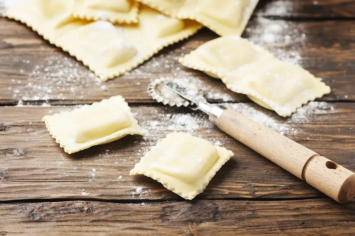 Świeże ciasto na ravioli, posypane mąką, leży na drewnianym blacie obok radełkowanego wałka.