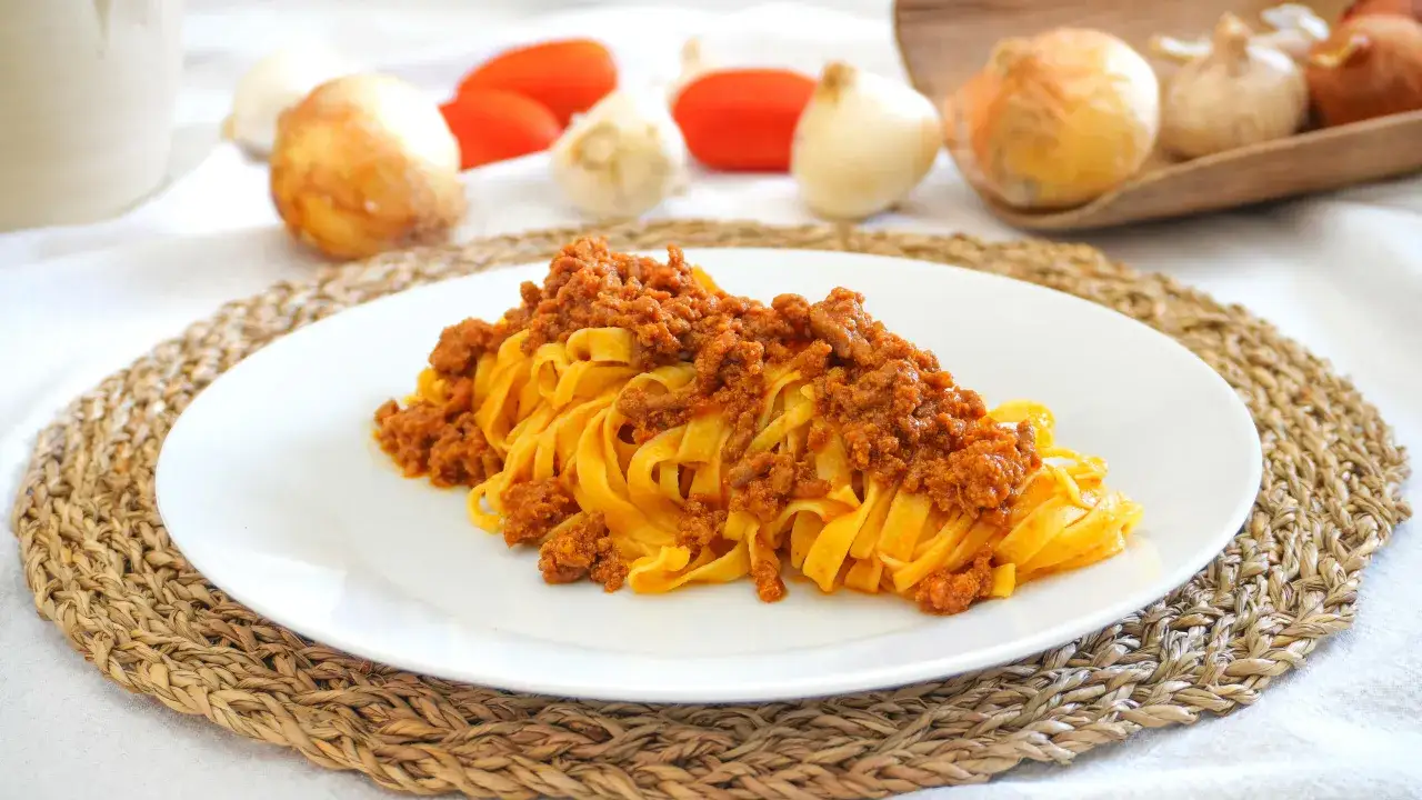 Un piatto invitante di tagliatelle al ragù, con ingredienti freschi come pomodori e cipolle sullo sfondo.