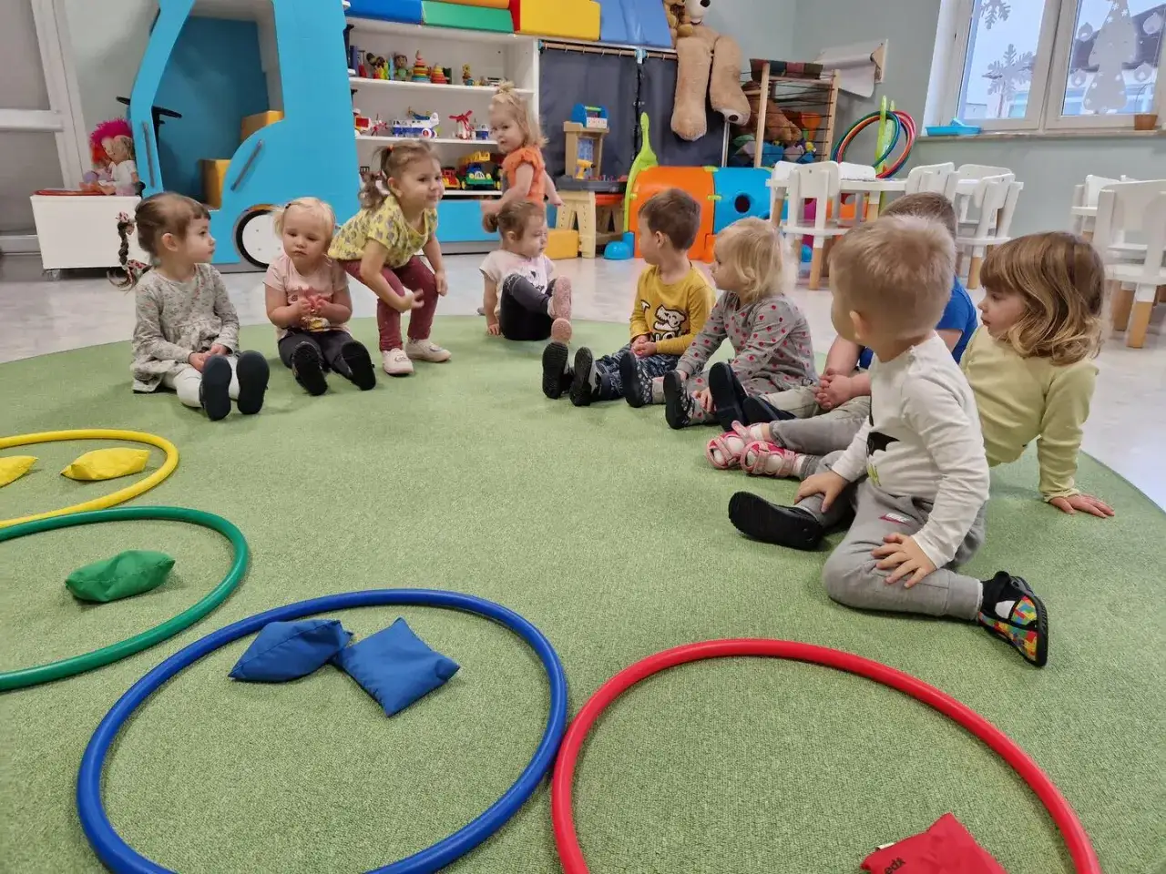 Dzieci siedzą na dywanie w żłobku, bawiąc się woreczkami i obręczami. Czy żłobek jest dobry dla dziecka? Tu widać radość i interakcję.