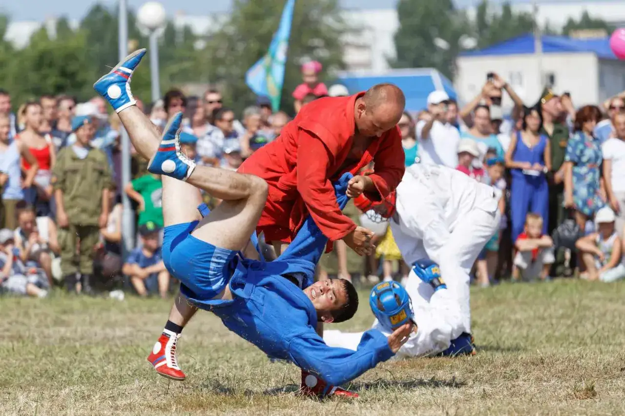 Tv&aring; brottare i combat sambo-dr&auml;kter k&auml;mpar p&aring; gr&auml;s. En i r&ouml;tt lyfter en i bl&aring;tt, som faller. Publik i bakgrunden.