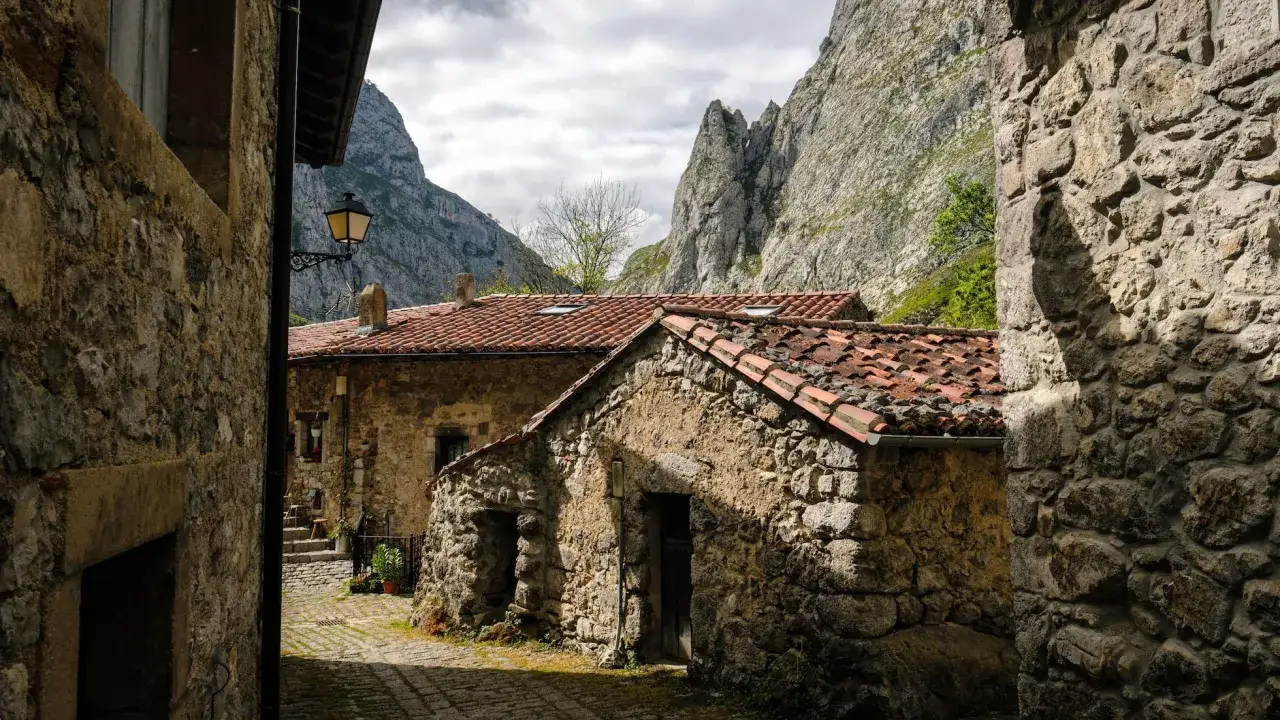 Calle empedrada entre casas de piedra con tejados rojos, bajo imponentes monta&ntilde;as. Un farol ilumina el camino, como un **ca&iacute;n de arriba** vigilando el pueblo.