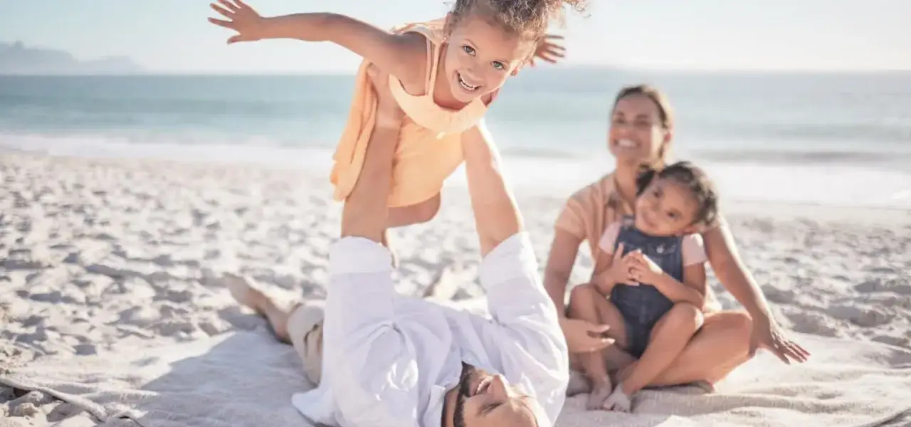 Radosny weekend z dzieckiem na plaży. Ojciec podrzuca córkę, mama z drugą córką siedzą obok.