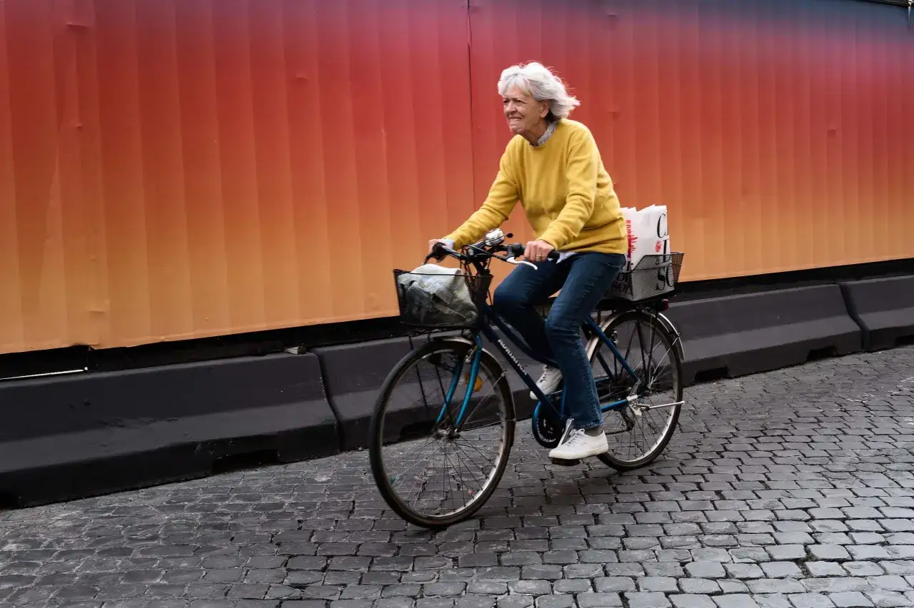 Donna sorridente in bicicletta con borse nel cestino, sullo sfondo un muro colorato. Leica Q3 43.