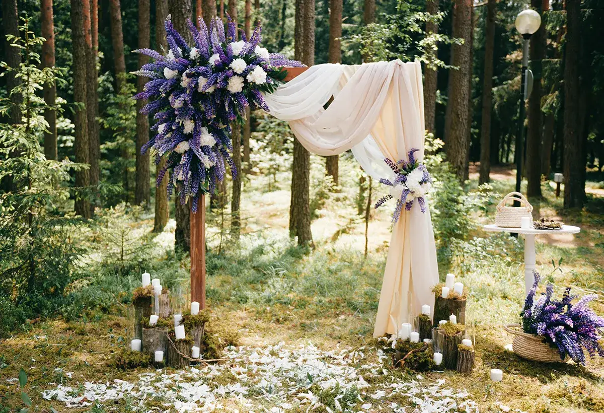 Un magnifique décor pour un mariage en forêt, avec une arche ornée de fleurs violettes et blanches, des bougies et des pétales au sol.