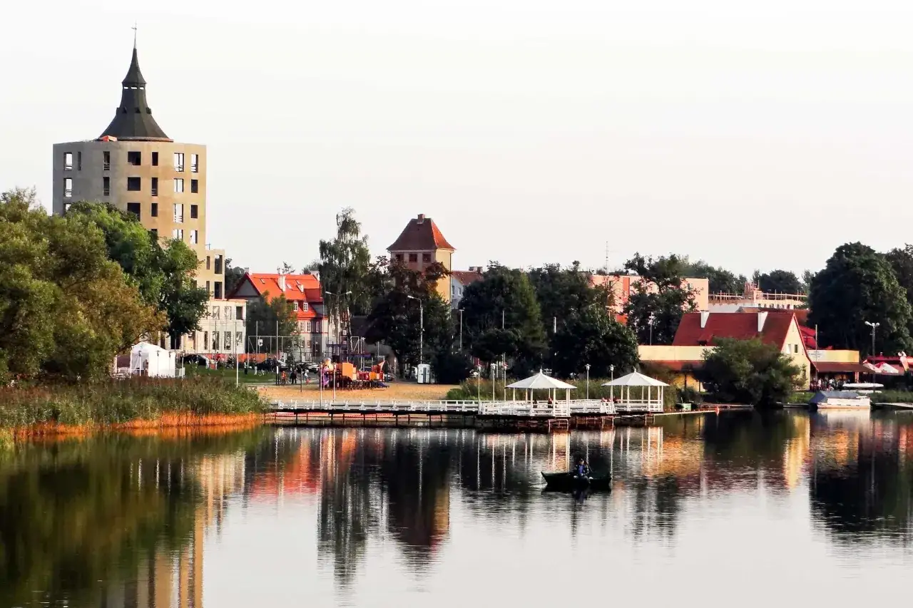 Widok na jezioro z łódką i zabudowaniami, w tym wieżą w budowie, tworzącymi malowniczy szczytno rynek.
