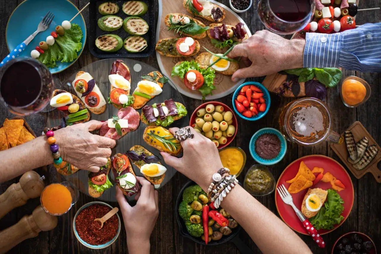 Una mesa llena de cosas t&iacute;picas de Espa&ntilde;a: tapas variadas, aceitunas, pimientos, patatas fritas y vino tinto.