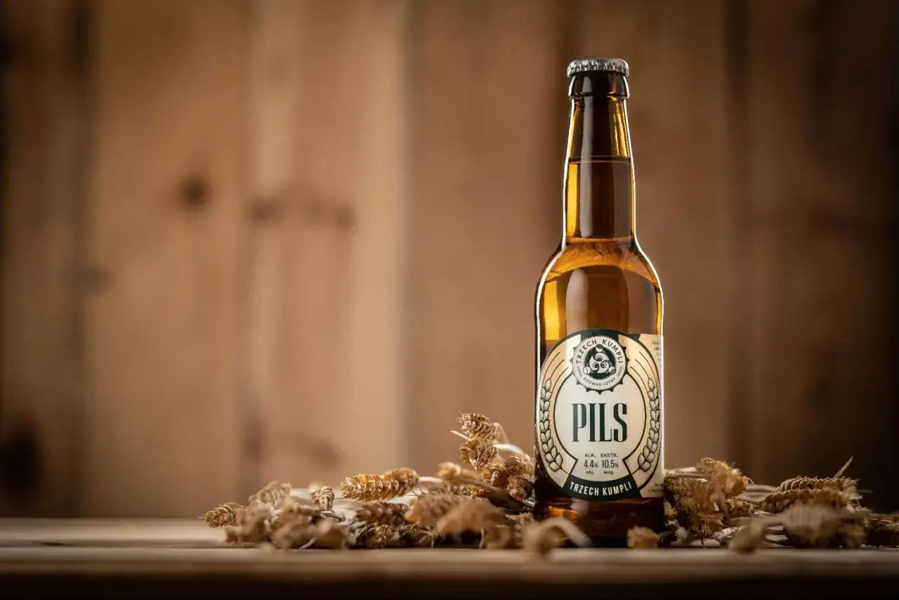 Butelka pils piwo od Trzech Kumpli, otoczona kłosami zbóż, na drewnianym tle.