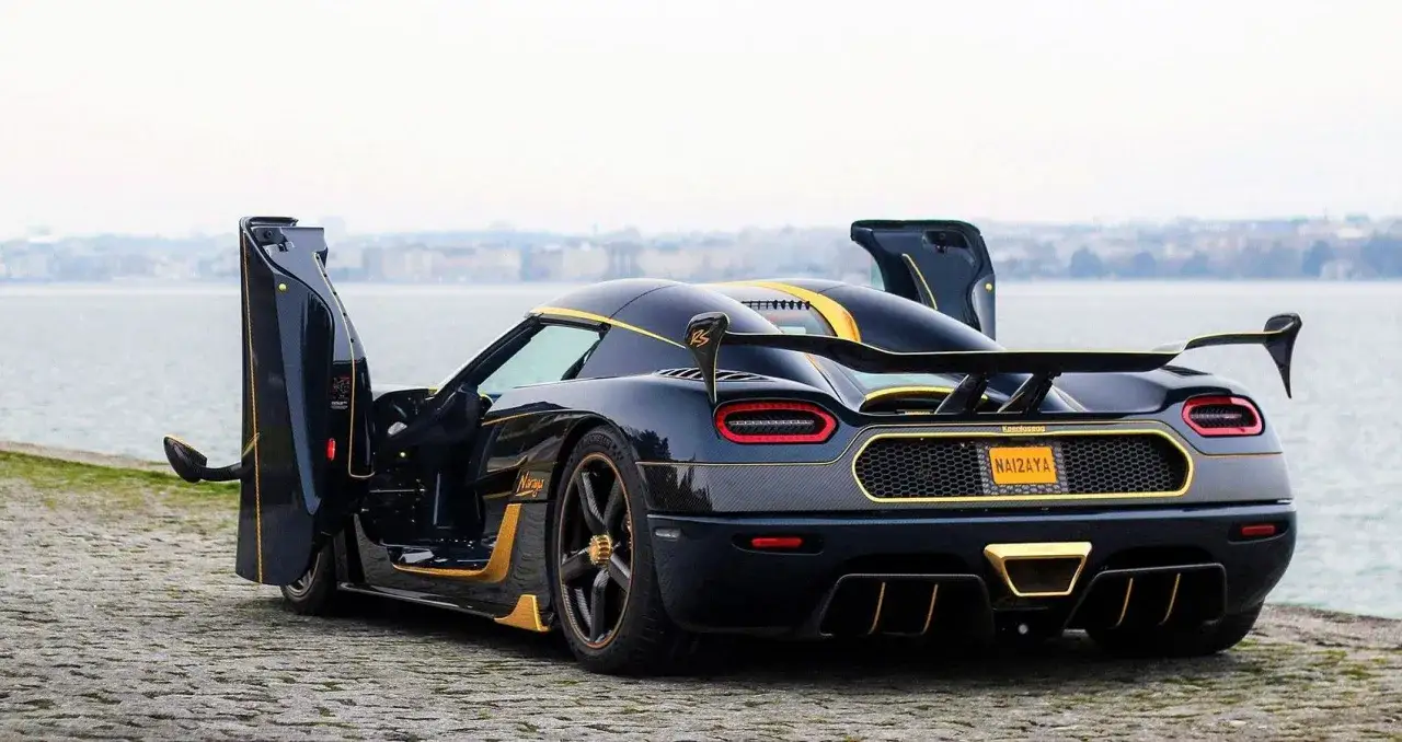 Czarny Koenigsegg Agera R ze złotymi akcentami i otwartymi drzwiami, zaparkowany nad wodą.