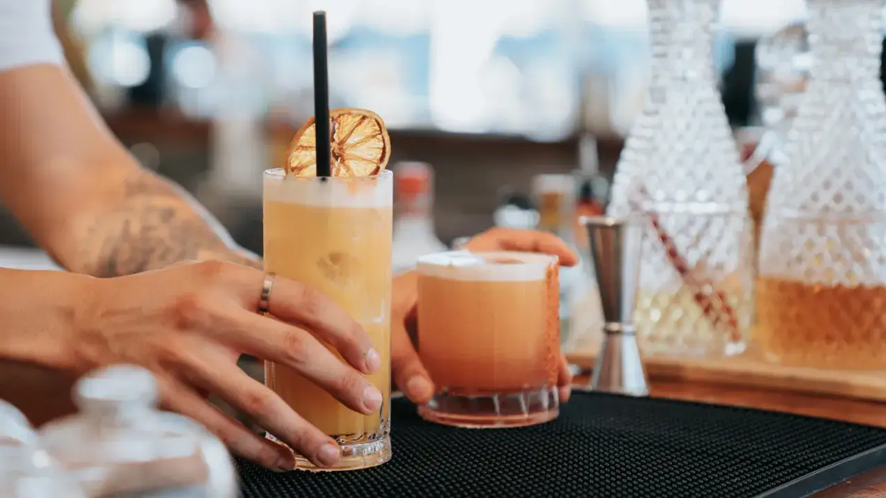 Dwie ręce barmana podają dwa klasyczne drinki: jeden wysoki, ż&oacute;łty z suszoną pomarańczą, drugi niższy, pomarańczowy.