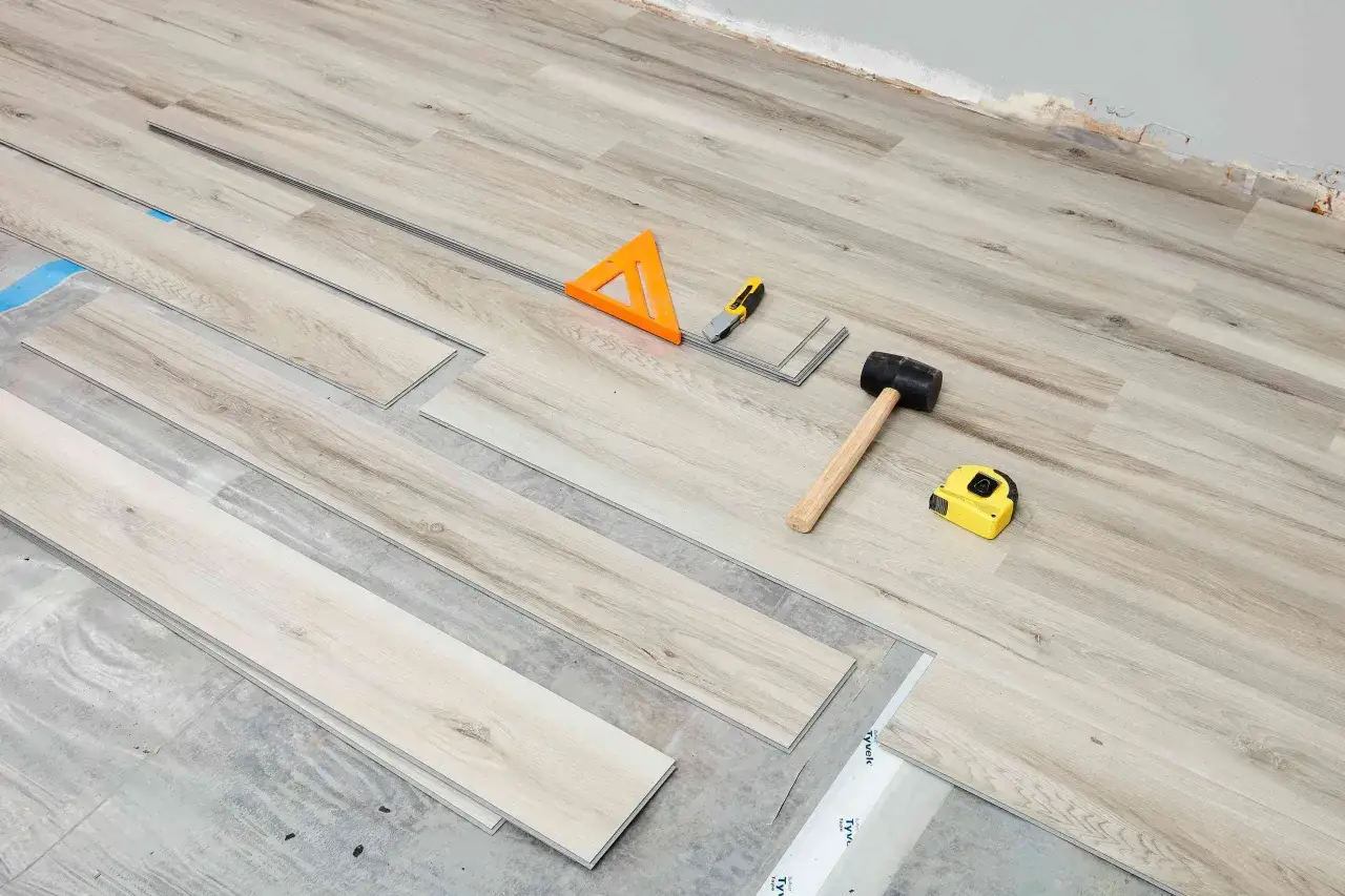 Tools for vinyl floor installation: planks, rubber mallet, utility knife, measuring tape, and a triangle square.