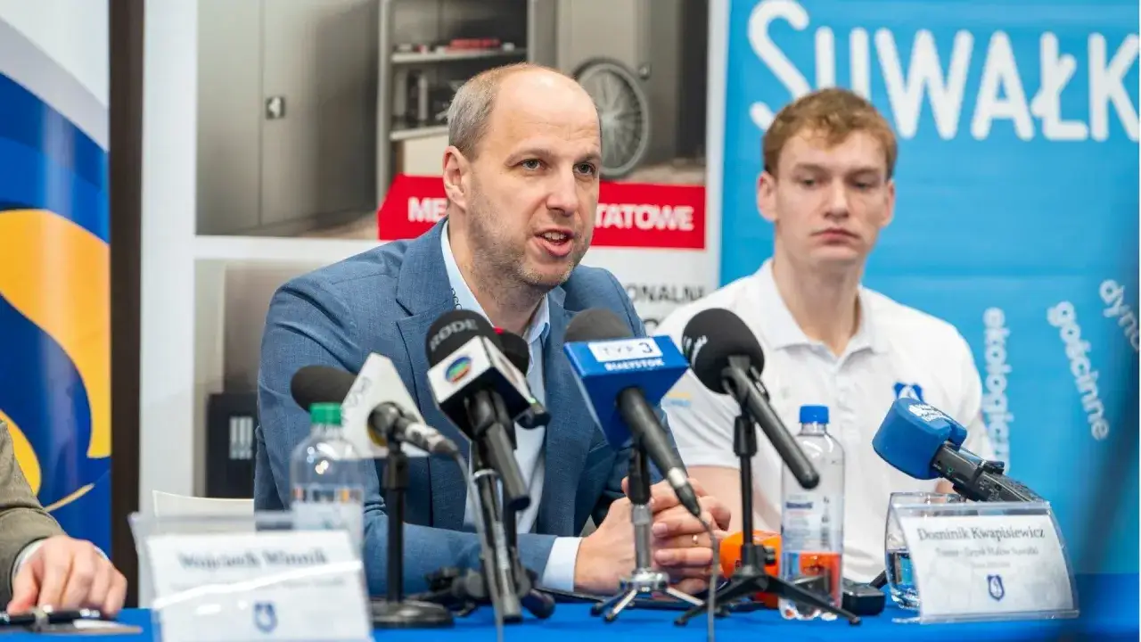 Dominik Kwapisiewicz, trener, przemawia do mikrofon&oacute;w podczas konferencji prasowej. Obok siedzi młody mężczyzna.