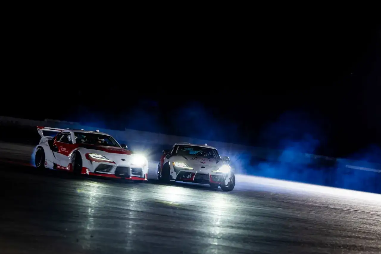 Dos coches Toyota Supra realizan un espectacular derrape coche en la noche, levantando humo azul y con sus faros iluminando la pista.