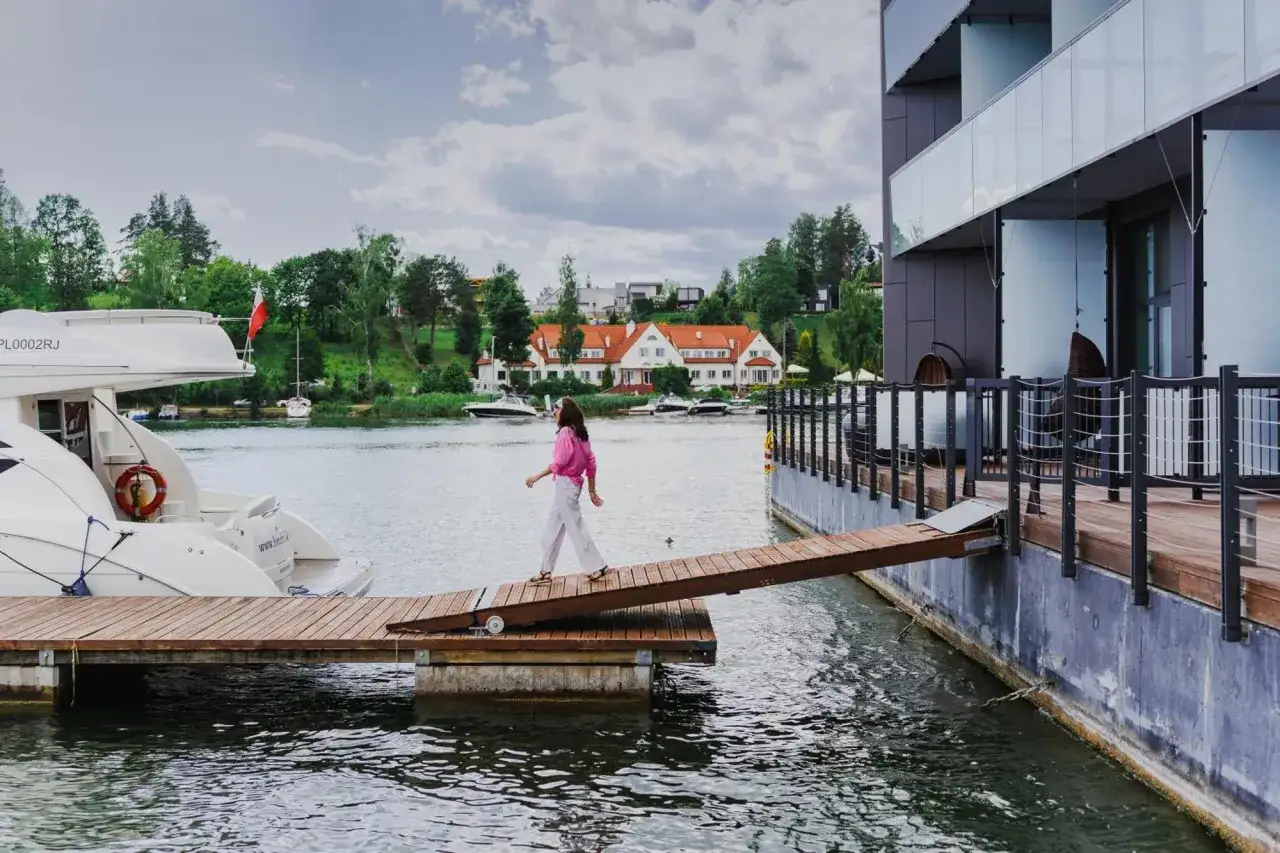 Kobieta idzie drewnianym pomostem do nowoczesnego hotelu Mikołajki Spa. W tle jezioro i malownicze domy.