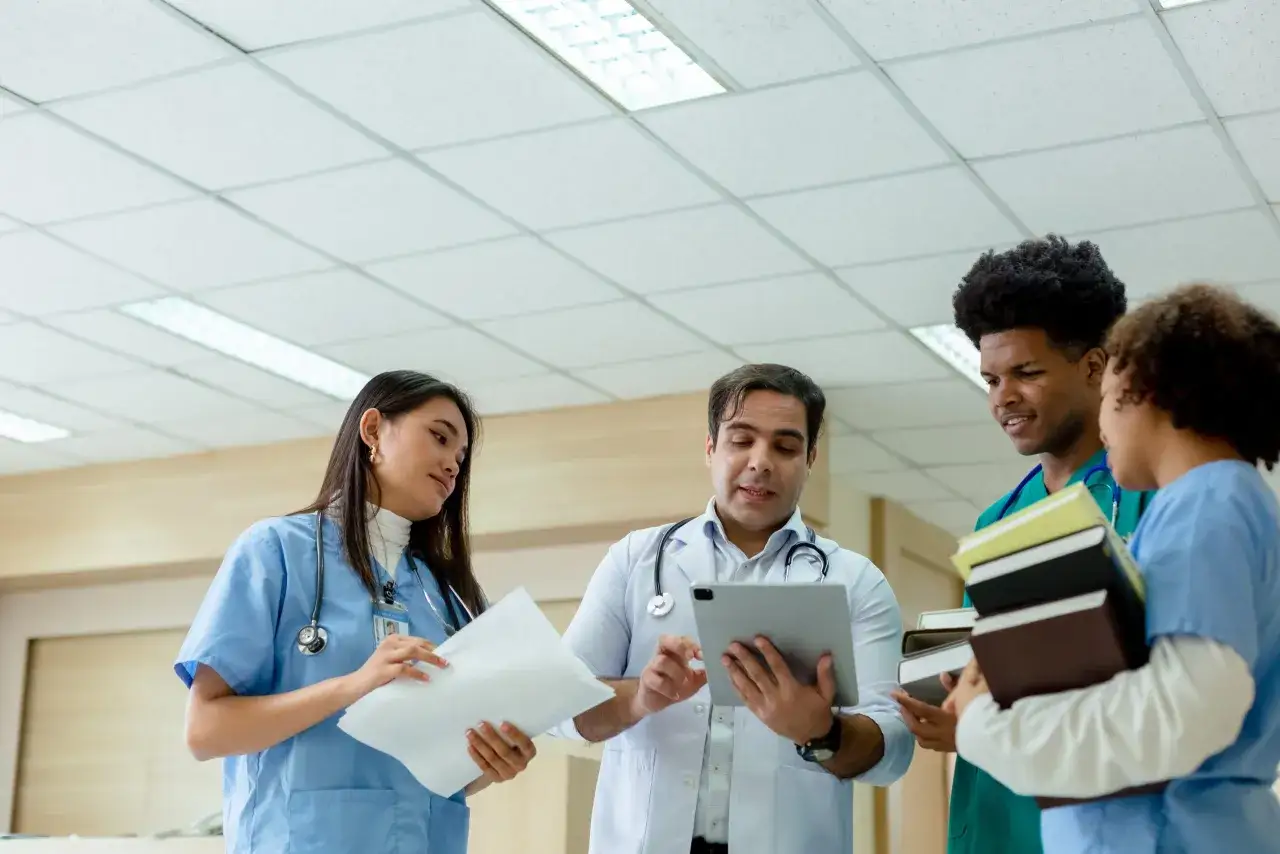Lekarze omawiają wyniki badań podczas studi&oacute;w dermatologicznych. Jeden z nich pokazuje coś na tablecie, a inni słuchają i trzymają książki.