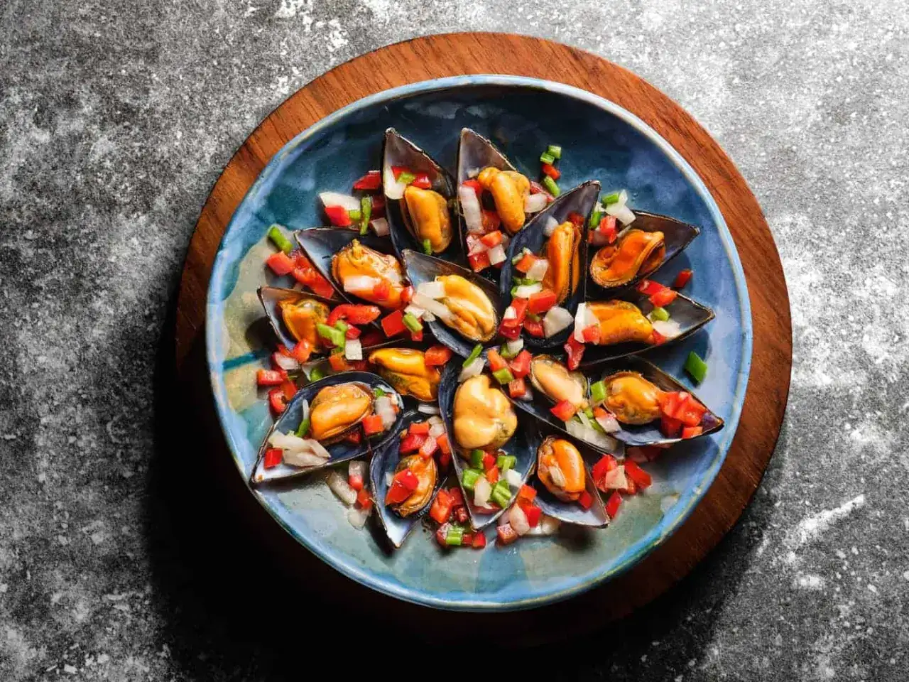Mejillones a la vinagreta, frescos y jugosos, servidos en un plato azul con pimientos y cebolla picados.