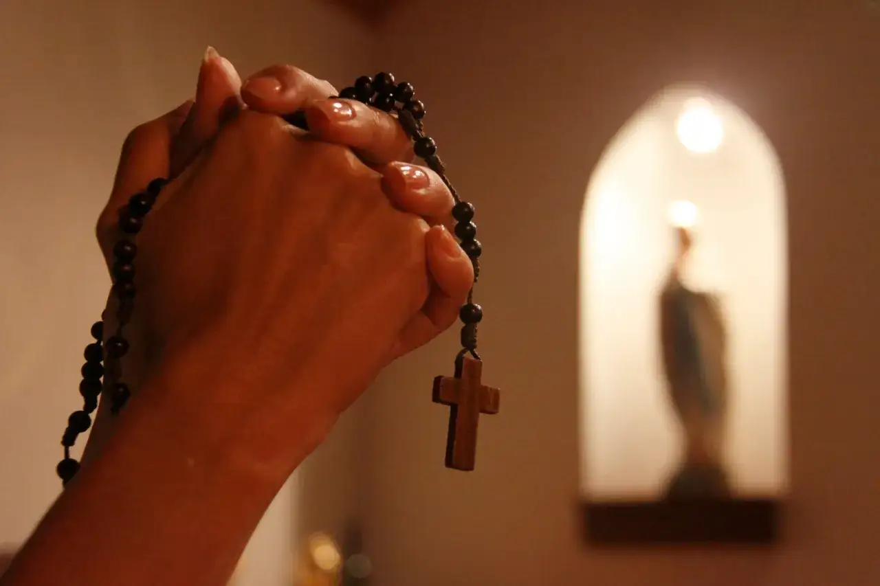 Dłonie z paciorkami r&oacute;żańca, w tle figura Matki Boskiej. Modlitwa do Maryi od spraw niemożliwych.