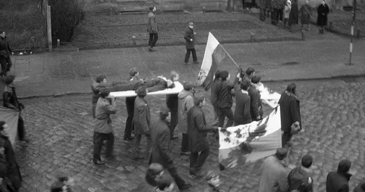 Grupa ludzi niesie nosze z ciałem, a obok powiewa biało-czerwona flaga. W tłumie widać transparent z napisem
