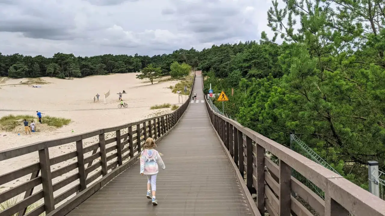 Dziewczynka idzie drewnianym molo przez wydmy w Dźwirzynie. W oddali widać plażowiczów i las.