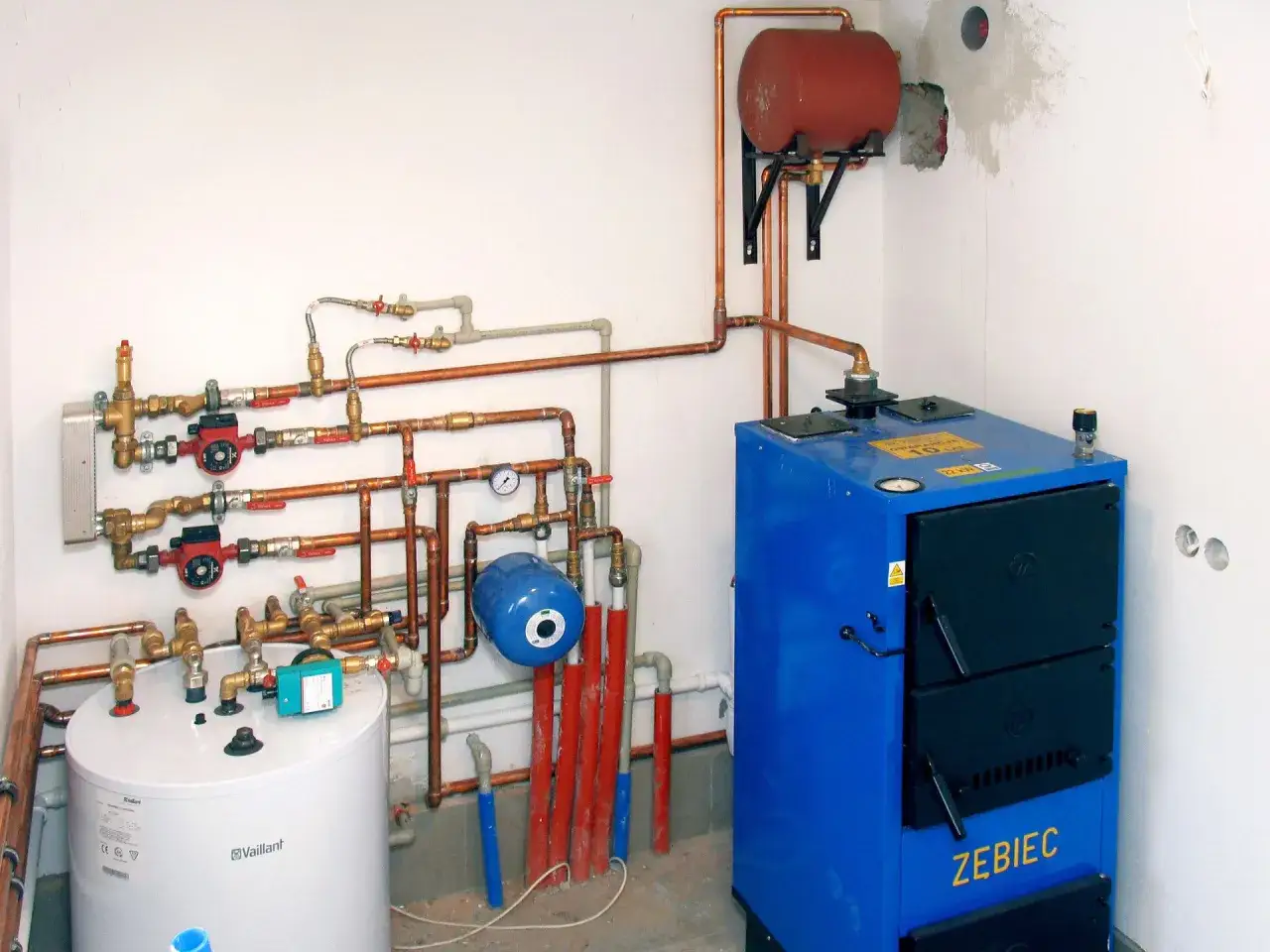Podłączenie pieca CO: niebieski piec Zebiec, biały zasobnik Vaillant, pompy, zawory i naczynie przeponowe tworzą instalację grzewczą.