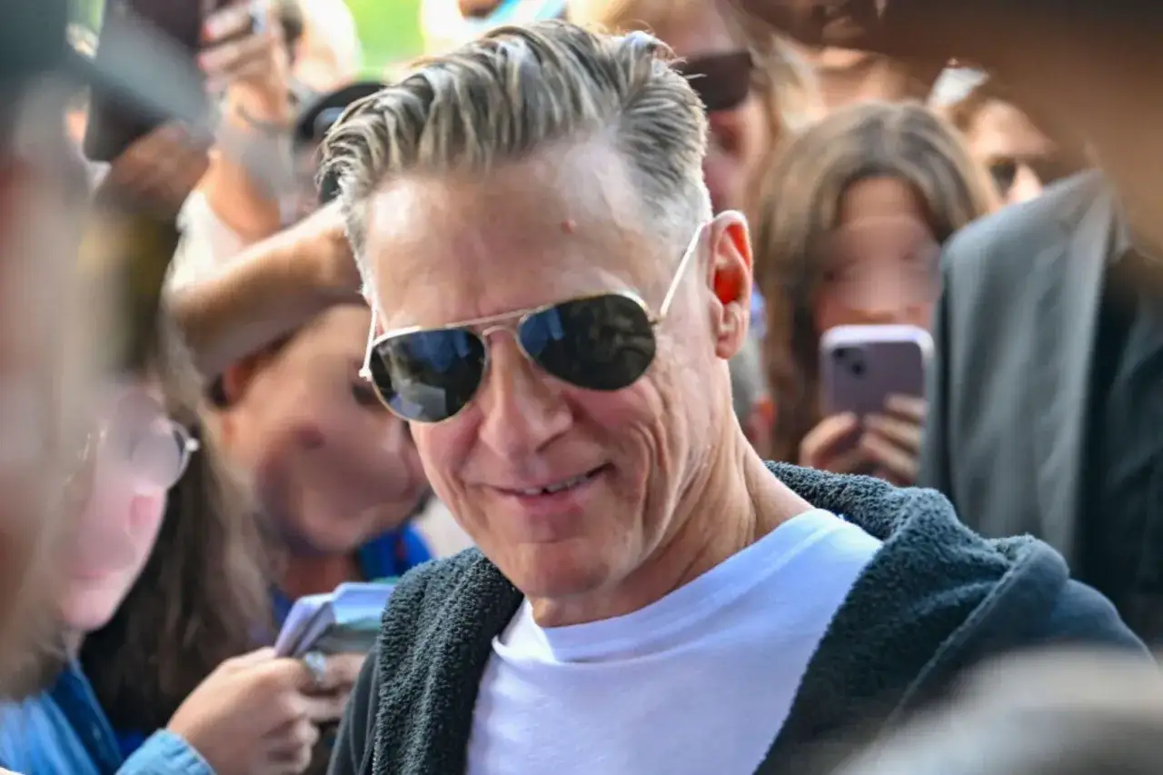 Bryan Adams w okularach przeciwsłonecznych, uśmiechnięty, otoczony fanami podczas koncertu.