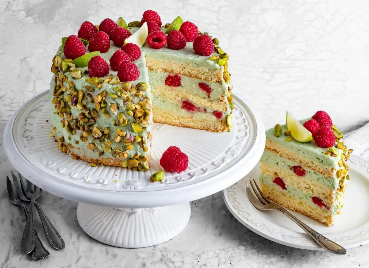 Pistacjowa torta z malinami i limonką, udekorowana orzechami. Kawałek ciasta na talerzyku obok.