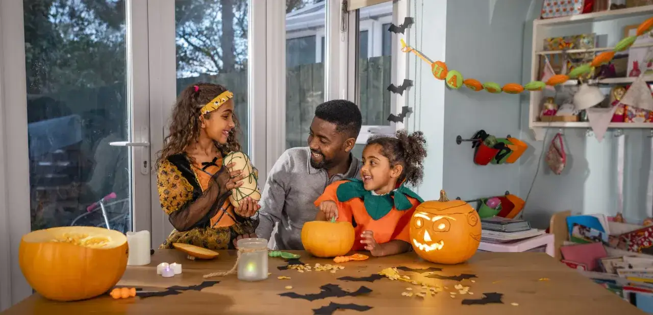 Un p&egrave;re et ses deux filles pr&eacute;parent leur **th&egrave;me soir&eacute;e halloween**. Citrouilles sculpt&eacute;es, costumes et d&eacute;corations cr&eacute;ent une ambiance festive.