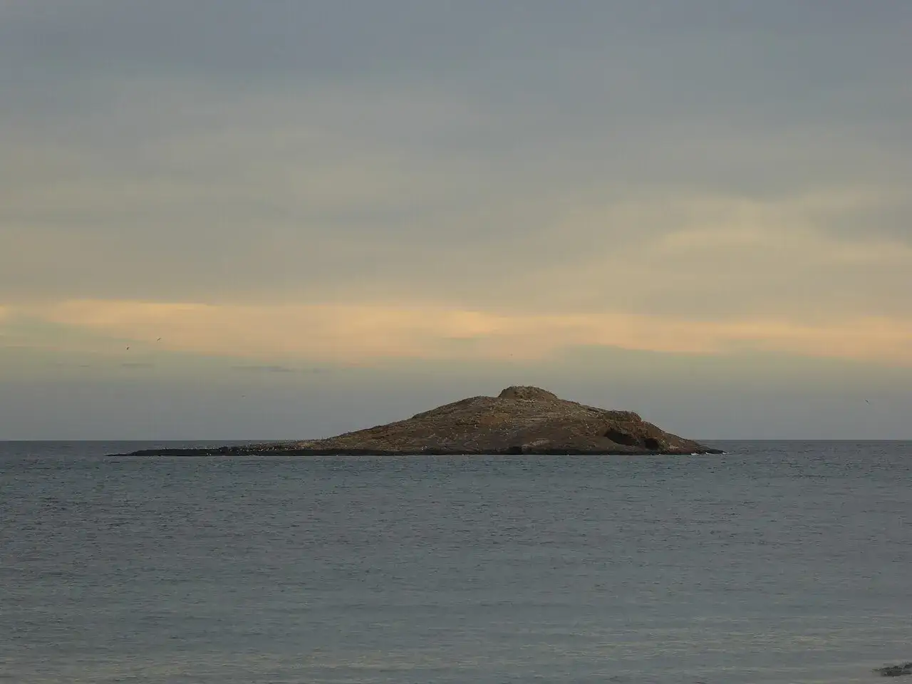 Isla de San Andrés, Almería, un islote rocoso solitario en el mar bajo un cielo nublado con tonos anaranjados.