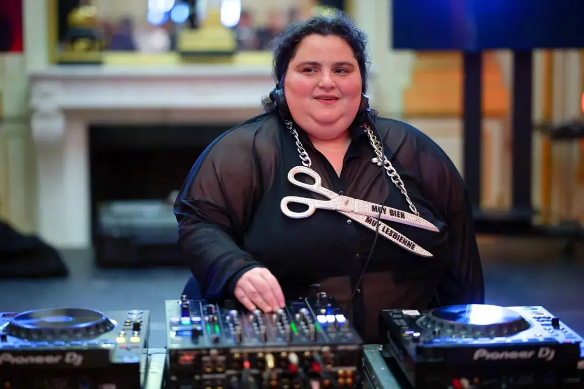 Une dj femme connue, aux platines, porte un collier en forme de ciseaux scintillants.