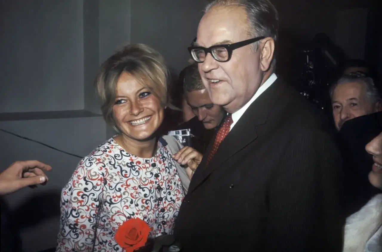 Monica Zetterlund, en glad maka, ler bredvid en man i kostym. En röd ros syns på hennes klänning.
