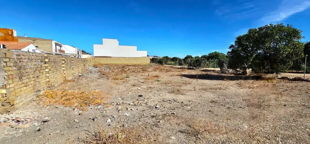 Un terreno es un bien inmueble con una pared de ladrillos, casas al fondo y &aacute;rboles.